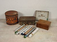 View of entire lot showing Club Chewing Tobacco large tin can, rusty metal tin, white Chesterfield cigarette tin, small wooden box, and six assorted tubes.