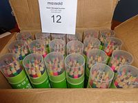 20 boxes of colored pencils arranged in a cardboard box. Each cylindrical green container has a transparent cap showing the sharpened colored pencils inside.