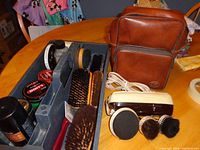 Wide shot of electric shoe polisher, multiple shoe brushes, shoe polish tins in a plastic caddy, and a brown carrying case on a wooden table.