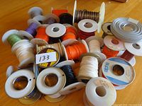 Multiple spools of hobby wire arranged on a wooden table. Variety of colors visible including white, purple, orange, black, and silver. Some spools have labels and different spool sizes.
