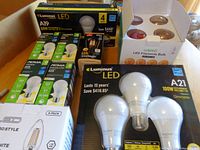 Photo showing multiple LED bulb boxes including Luminus A19 60W 4-pack, NOMA A19 single packs, LED Vintage filament bulb pack and Luminus A21 100W 3-pack on a wooden table.