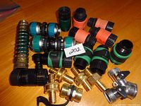 Photo of assorted garden hose connectors, including green and orange plastic connectors and couplers, brass fittings, and small metal parts spread on a wooden surface.