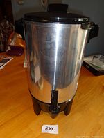 Front side view of stainless steel West Bend coffee machine with black plastic base, spout, and locking lid, placed on a wooden table.