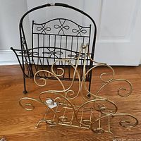 View of two metal magazine racks together on a wooden floor in front of a white door. Black rack upright with handle, gold rack in front with scroll design.