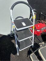 3 tier white metal step stool with black slip-resistant steps, folded in open position outside in sunlight.
