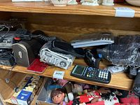 Photo showing several electronics including two scientific calculators, a compact scale, a camcorder, and a device wrapped in plastic identified as a track radio CD player.