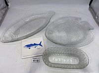 View of two clear glass fish-shaped platters and one bluefish ceramic trivet. Platters have textured scale patterns and fins, trivet shows bluefish illustration.