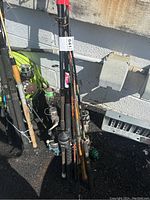 Photo showing multiple fishing rods and reels leaning against a wall outside. Various handle styles and reel types visible.