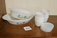 Two white Glasbake casseroles with green daisy floral pattern stacked and placed next to six white custard/storage cups and six plastic lids on a wooden surface.