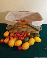 Large woven picnic basket with white cloth interior and metal handles, with assorted faux fruit displayed in and around the basket on a dark green surface.