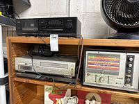 Photo showing the wooden shelving holding several electronics including a TEAC cassette deck with tuner, a Penske automotive analyzer, a Panasonic VHS recorder, and a Samsung DVD player. Also visible is a black remote control device.