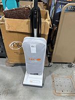 White upright Lux Lite vacuum with gray dust bag and black handle standing on concrete floor in front of cardboard box.