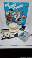Photo showing all items including Three Stooges record, Lenox cup/saucer, cufflinks box, travel razor, metal cases, key fob, and additional accessories.