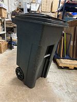Side angle showing the tall rectangular black plastic trash can with wheels and side handles in a warehouse environment.