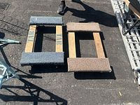 Two 4-wheel roller dollies side by side on asphalt, one blue carpeted and one beige carpeted.
