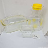 Three clear glass kitchen items including a lemonade pitcher with yellow lid, a square refrigerator dish with lid, and a loaf dish with lid.