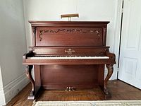 Front view of the antique Heintzman upright grand piano showing carved woodwork, keyboard, pedals, and piano light.