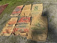 Seven worn burlap sacks arranged on the ground, showing different faded printed logos and various sizes.