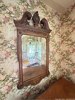 Full view of the carved wooden mirror hanging on floral wallpapered wall showing reflection of room.