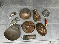 Photo showing early copper kitchen moulds and utensils arranged on metal surface including perforated pan, fruit and fish shaped moulds, sieve and other tools