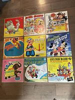 Top-down photo showing all nine vinyl record covers arranged in a 3x3 grid on the floor. Cartoon characters featured include Bugs Bunny, Mighty Mouse, Popeye, Mickey Mouse, Bozo Circus, Robin Hood with Top Cat, Quick Draw McGraw, and Huckleberry Hound.