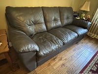 Front angle view of the brown leather couch showing three padded seat and back cushions, on wooden floor near a side table with lamp.