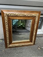 Front view of the mirror in its entirety showing the wooden frame with gold ornate detailing and clear mirror surface.