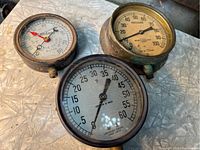 Four vintage pressure gauges on a textured surface, showing visible age, rust, and damage to glass faces including one missing glass and one cracked.