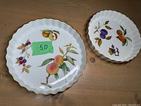 Two Royal Worcester ceramic tart dishes with scalloped gold-trimmed edges on a wooden surface. The larger dish features peaches, blackberries, and apples. The smaller dish features plums and oranges.