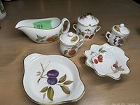Top view of six Royal Worcester porcelain cookware and serving pieces with painted fruit designs on white porcelain with gold trim.