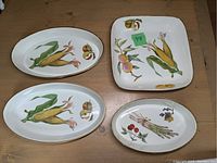 Four Royal Worcester ceramic baking dishes displayed on a wood surface. Designs include corn, peaches, asparagus, and cherries with gold trim.