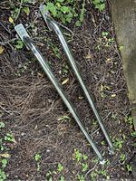 Two chrome truck bed rails placed on the ground with leaves and pine needles around. Rails have mounting brackets attached on ends.