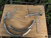 Two bicycle fenders and wire basket laid out on a wooden surface showing chrome finish and mounting parts.