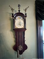 Full view of clock mounted on wall, showing wooden case, dial, weights and chains