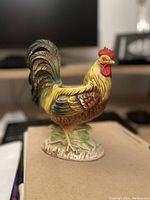 Front view of the hand painted pottery rooster figurine showing detailed feathers, yellow and brown body, red comb and wattle, standing on a leaf-shaped base.