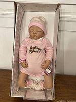 Full view of Baby Madison doll in its original box wearing pink onesie, hat, and booties, including certificate of authenticity and bracelet in the box.