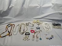 Overview photo showing all jewelry items laid out, including various fashion necklaces, earrings, and a bracelet, some with tags attached, displayed on a white cloth background.