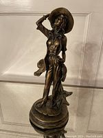 Front view of bronze sculpture depicting a woman in a hat with detailed clothing and expressive pose on a circular base.