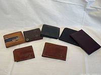 Photo showing seven assorted men's leather wallets and billfolds arranged on a light cloth surface, varying in colors and styles.
