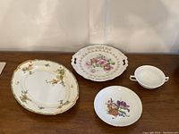 Four items displayed: large oval floral platter with gold trim, decorative handled platter with pink rose design and gold German text, white bowl with purple and orange flowers, and a small white handled bowl.
