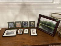 Nine framed artworks including carved relief portraits, landscape photography, and architectural prints arranged on a wooden table.