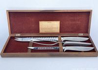 Opened wooden box with red velvet lining displaying two carving knives and one carving fork, all with stainless steel ergonomic handles and blades, arranged inside the box. An informational label about the Gerber Legendary series is attached to the inside lid of the box.