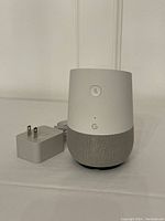 Google Home smart speaker with original power adapter on white surface showing top mute button and Google 'G' logo.