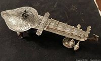Top angle showing intricate floral engravings, tuning pegs, strings, and overall shape of the miniature sitar container.