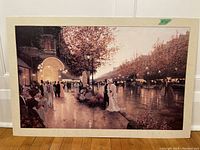 Full view of the print showing the elegant evening street scene with figures, illuminated boulevard, trees, and warm autumn tones, unframed, laid against a white door.
