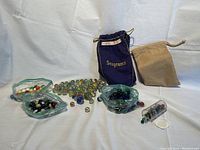 Top view of collection including several groups of assorted vintage glass marbles arranged on a white surface. Two drawstring bags (a blue Seagrams brand velvet bag and a plain tan bag) are visible behind marbles. Some marbles are loose, others are in plastic zippered bags.