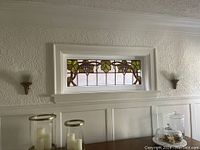 Horizontal stained glass window panel with grapevine motif installed in white wooden frame above wall paneling with candle holders and table below.