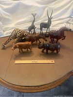 Seven wooden animal carvings displayed on table with ruler for scale, showing various animals including giraffe, antelope, horse, cow, and zebra.
