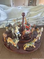 Image showing multiple ceramic carousel animal figures placed on a circular wooden base with a central carved handle. Animals include horses, a giraffe, lion, camel, zebra, leopard. Figures are decorated with painted saddles and bridles.