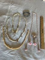 Photo of all jewelry items laid on a white cloth including 4 necklaces, black bead bracelet, floral pin, floral earrings, and a ruler for scale.